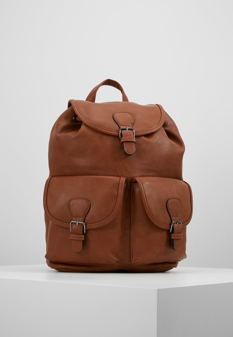 Sac à dos en cuir marron avec deux poches avant à boucle et un rabat supérieur fermé par une boucle, posé sur un socle blanc sur un fond gris.