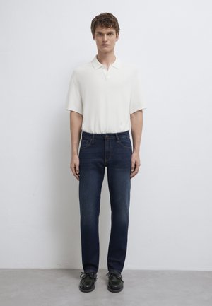 Junger Mann steht an einer weißen Wand, trägt ein weißes kurzärmeliges Poloshirt, dunkelblaue Jeans und schwarze Schuhe.