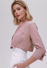 Veste courte rose pâle avec une texture lisse, un design structuré et des manches trois-quarts avec des accents en nœud aux poignets.