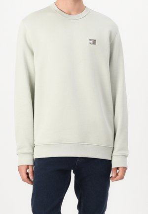Mężczyzna ubrany w jasnoszary sweter z okrągłym dekoltem z małym logo "Tommy Jeans" na lewej piersi oraz ciemnoniebieskie dżinsy.