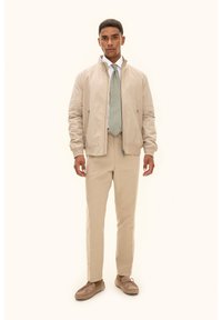 Beige Bomberjacke, weißes Hemd, gemusterte grüne Krawatte, beige Hose und hellbraune Schuhe; glatter Stoff und lässige Passform.