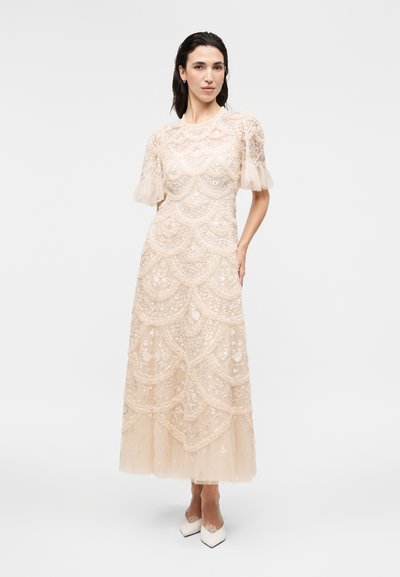 Γυναίκα με μπεζ δαντελένιο μίντι φόρεμα με κυματιστά σχέδια, διάφανα φουσκωτά μανίκια και λευκές μυτερές γόβες, στέκεται με το ένα χέρι στη μέση.