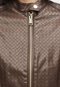 Καφέ δερμάτινο μπουφάν με καπιτονέ υφή, με χρυστό φερμουάρ και κούμπωμα με κουμπιά στο γιακά. Σχεδιασμένο με εφαρμοστή σιλουέτα.