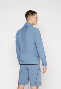 Lys blå treningsjakke med teksturert bakpanel, lange ermer og glidelås. Kombinert med matchende shorts med sidelommer.