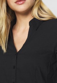 Zwarte blouse met een V-hals en kraag, voorzien van knoopdetails. Gladde stofstructuur en eenvoudig ontwerp. Geen aanvullende patronen zichtbaar.
