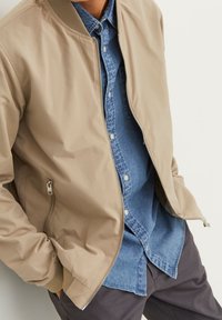 Beige bomberjacka med ribbad krage, dragkedjestängning och mjuk, slät yta, lager på lager över en blå denimskjorta.