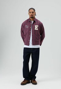 Rood- en marineblauw geruit overhemd met knopen, met een geborduurde letter "E" en tekst; gedragen over een wit T-shirt. Gecombineerd met donkere jeans en bruine laarzen.