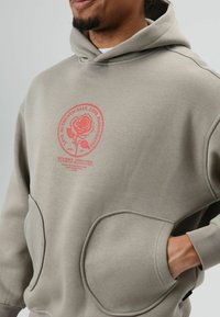 Sweat à capuche gris clair en tissu doux, orné d'un grand motif de rose rouge, avec de grandes poches kangourou. Texture lisse.