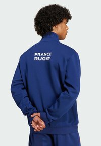 Marineblaue Rugbyjacke mit hohem Kragen, mit "FRANCE RUGBY" in Weiß auf dem Rücken. Glatte Textur und lockere Passform.