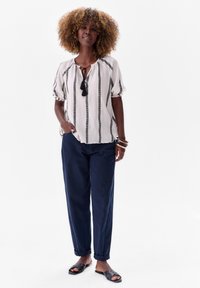 Witte blouse met zwarte verticale strepen, korte pofmouwen en een strik bij de hals. Marineblauwe, comfortabele broek. Zwarte instapsandalen.