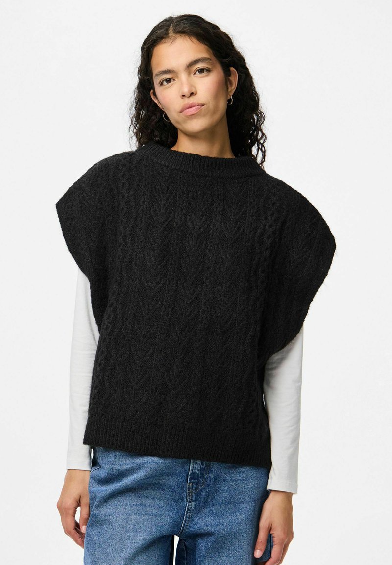 Svart stickad tröja med en texturerad kabelmönster, lös passform, korta ärmar och hög halsringning, bärs över en vit långärmad skjorta.