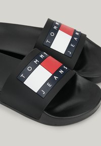 Sandales à glissière noires avec une large bride, affichant le logo Tommy Jeans en rouge, blanc et bleu marine sur le devant de chaque sandale.