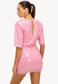 Robe en sequins rose avec des manches courtes, forme ajustée, détail en trou de serrure au dos et surface texturée. Accentuée par une boucle d'oreille en forme de soleil doré.