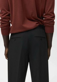 Osoba w brązowej koszuli z długim rękawem, włożonej w czarne spodnie z wysokim stanem, z zapiętymi tylnymi kieszeniami, pokazana od pasa w dół.