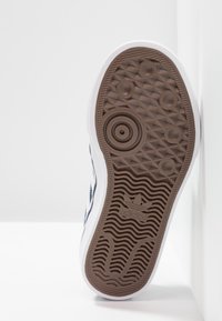 Vit sneakersula med en brun gummitr tread som har cirkulära och zigzag-mönster, präglad logotyp och texturerad yta för grepp.