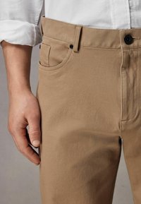 Beżowe chinosy z teksturowanej tkaniny, z boczną kieszenią, czarnym zapięciem na guzik i przeszyciami wzdłuż pasa i kieszeni.