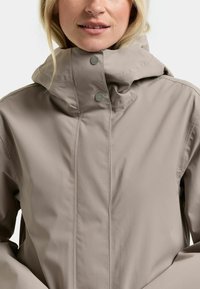 Vaaleanbeige vedenpitävä takki, jossa on korkea kaulus, napsautettavat kiinnikkeet ja sileä, mattapintainen viimeistely. Painopiste toiminnallisuudessa ja mukavuudessa.