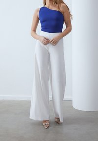 Blå enaxlad bodysuit i kombination med vita vida byxor. Skorna har genomskinliga remmar med dekorationer. Släta texturer.