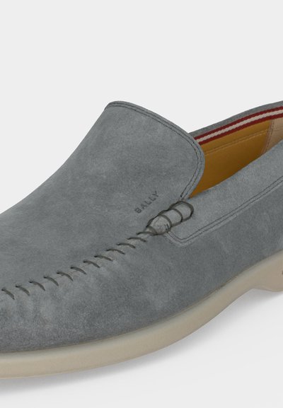 Γκρι σαμουά μοκασίνι χωρίς κορδόνια με ραμμένα σχέδια, μπεζ σόλα και κόκκινο-άσπρη ριγέ εσωτερική επένδυση, με το λογότυπο "BALLY" κοντά στο μπροστινό μέρος.