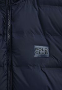 Granatowa pikowana kurtka z przednim zamkiem błyskawicznym i naszywką w kształcie prostokąta z napisem "POLO CLUB" na piersi.