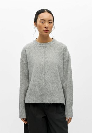 Femme aux cheveux foncés attachés porte un pull en maille gris clair et un pantalon noir, debout devant un fond blanc uni.
