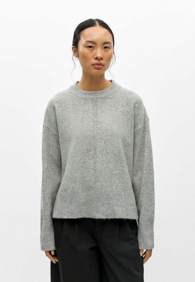 Femme aux cheveux foncés attachés porte un pull en maille gris clair et un pantalon noir, debout devant un fond blanc uni.
