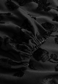 Czarna tkanina z kwiatowym wzorem, fakturowana aksamitnymi akcentami. Materiał ma miękką strukturę i subtelny połysk. Obecne detale w formie marszczeń.