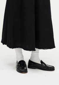 Czarne skórzane mokasyny z marszczonym zdobieniem, noszone z białymi prążkowanymi skarpetkami oraz czarną spódnicą o długości do połowy łydki z postrzępionym brzegiem.