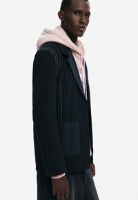 Chaqueta de mezclilla con costuras en contraste, un bolsillo y acabado negro, superpuesta sobre una sudadera con capucha de color rosa claro. Tejido texturizado.
