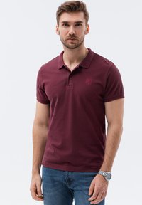 Burgundská polo košile s krátkým rukávem, žebrovaným límcem a vyšitým logem. V kombinaci s modrými džíny a stříbrnými hodinkami.