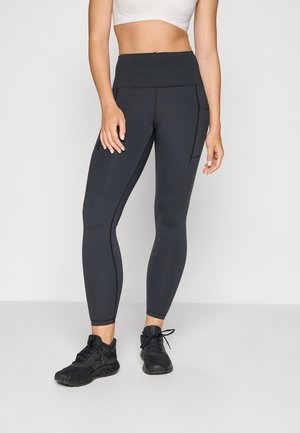 Leggings gris foncé taille haute en tissu lisse et extensible. Comprend des coutures le long des jambes et une poche latérale. Associés à des chaussures de sport noires.