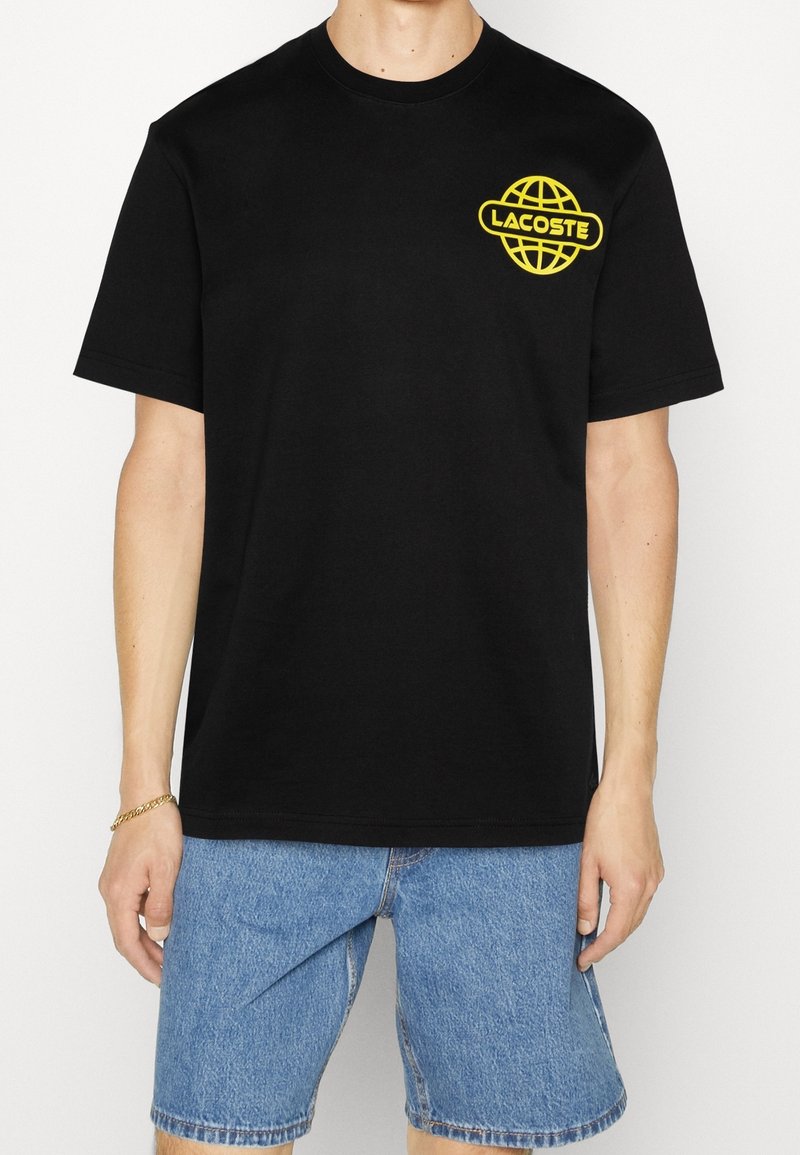 Black cotton t-shirt featuring a yellow globe logo with "LACOSTE" text on the left chest, short sleeves, and a relaxed fit.