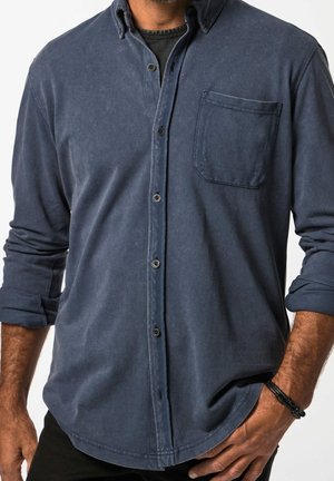 Homme portant une chemise bleu foncé à boutons avec les manches retroussées et un bracelet noir au poignet gauche, main dans la poche.