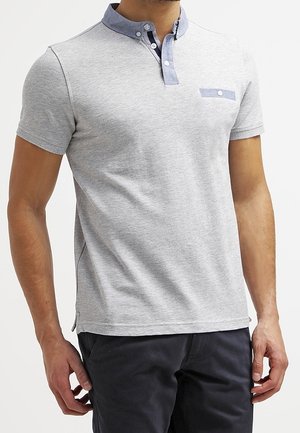 Mężczyzna ubrany w szary polo z krótkim rękawem, niebieskim kołnierzykiem na guziki i kieszonką na piersi, w ciemnych spodniach, stojący na jednolitym tle.