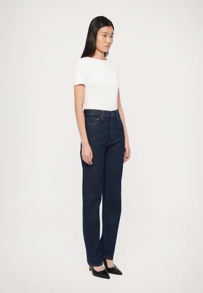 Tmavomodré džínsy s rovnými nohavicami a vysokým pásom, doplnené priliehavým bielym tričkom s krátkym rukávom a čiernymi lodičkami so špicatou špičkou.