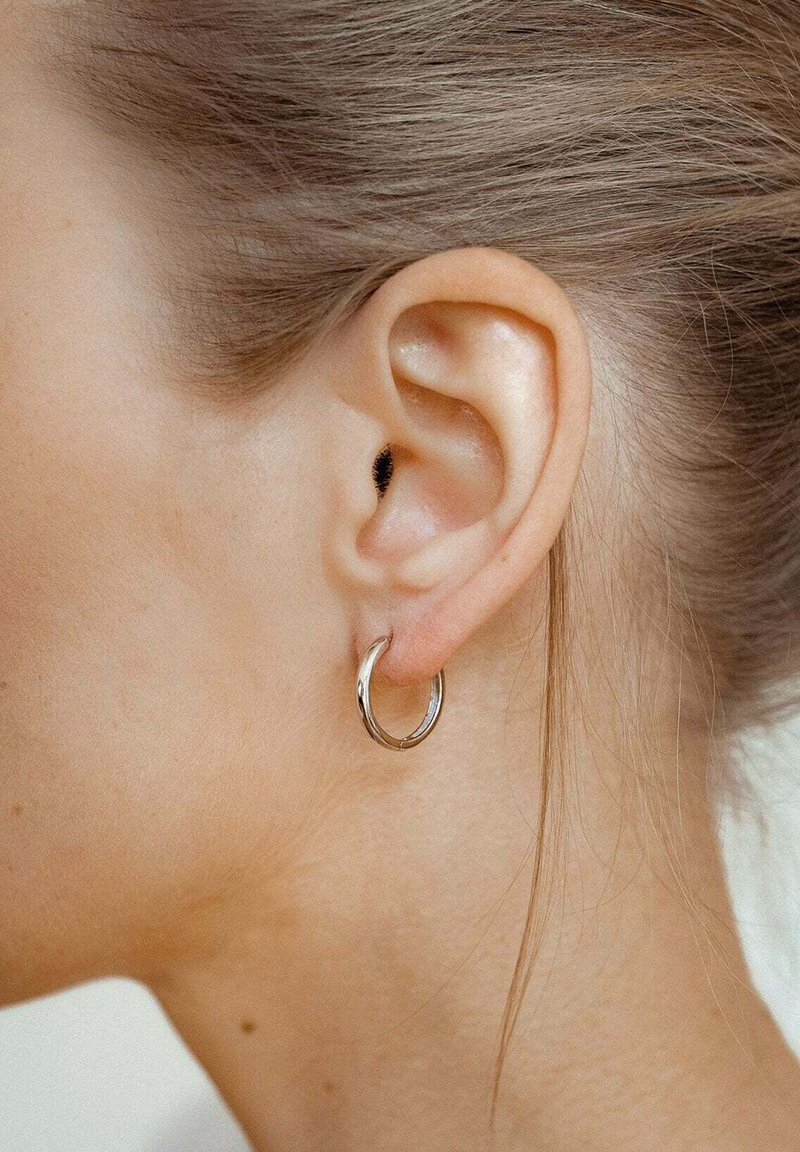 Silberne Creolen, klein und rund, mit einer glänzenden Oberfläche, getragen im gestochenen Ohr, das vor hellbraunem Haar zur Geltung kommt, mit einem schlichten Design.