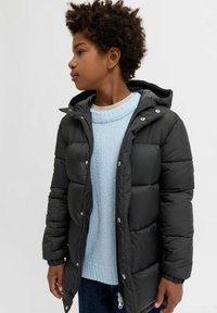 Melnā pūkainā jaka ar kapuci, ar rāvējslēdzēju un pogām. Uzvilkta virs gaiši zilas struktūrētas džemperis un tumšiem džinsiem.