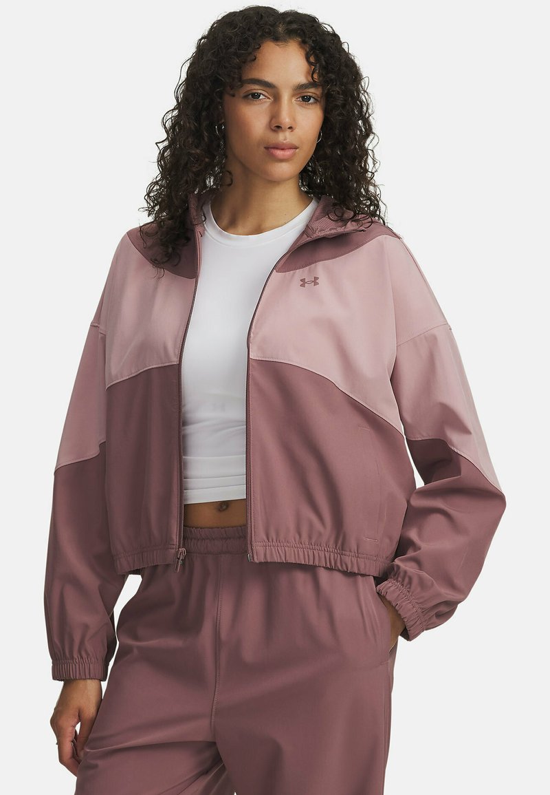 Veste coupe-vent rose courte avec un zip à l'avant, des panneaux contrastants, des poignets élastiques et une taille élastique, associée à un haut blanc.