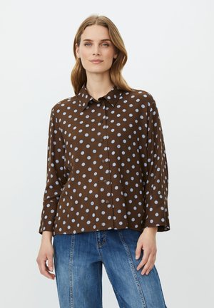 Kvinde med lyst brunt hår iført en brun langærmet skjorte med hvide polkaprikker og blå jeans, stående foran en ensfarvet baggrund.