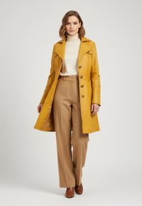 Yellow leather trench coat with a belted waist, buttoned front, and wide lapels, paired with beige trousers and brown shoes.