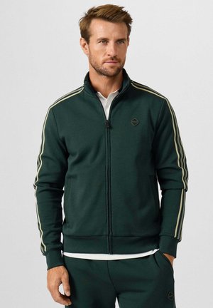 Man draagt een donkergroene jas met rits en bijpassende broek met lichte strepen op de mouwen, staand voor een effen achtergrond.