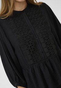 Black button-up blouse with a textured, embroidered center panel and gathered hem. Features long sleeves and a rounded neckline.