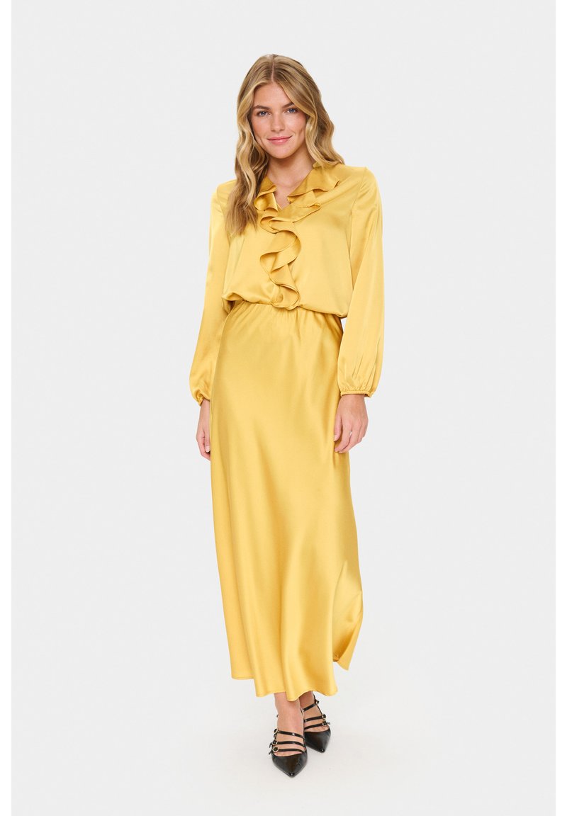 Donna che indossa un lungo abito di raso giallo con scollatura arricciata e maniche lunghe, abbinato a tacchi neri a punta con cinghie.