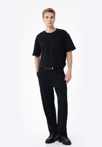 Czarna koszulka z krótkim rękawem w połączeniu z czarnymi spodniami, z paskiem i czarnymi butami, na tle jasnego, jednolitego tła.