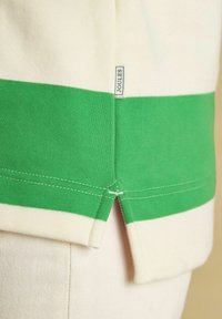 Nahaufnahme eines cremefarbenen und grünen gestreiften Stoffes mit einem Joules-Markenetikett und einem seitlichen Saumschlitz an einem Kleidungsstück.