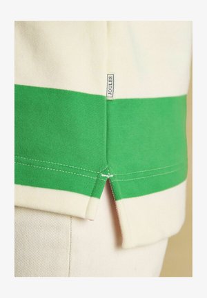 Kreemja ja rohelist triibulist kangast lähivõte, millel on Joulesi kaubamärgi silt ja küljeääre avaus rõival.