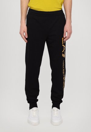 Homme portant un pantalon de survêtement noir avec le logo "EA7 Emporio Armani" doré sur la jambe droite, associé à des baskets blanches et un haut jaune.
