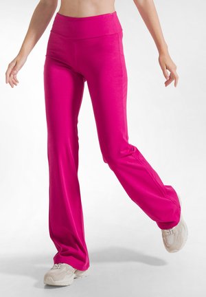 Vrouw wearing felroze flare broek en witte sneakers, staand met één been omhoog tegen een effen achtergrond.