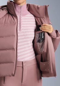 Jastućasta ružičasta jakna s visokim ovratnikom, prednjim patentnim zatvaračem, ispod koje je teksturirani pleteni džemper, te džep za pametni telefon s prozirnim poklopcem.