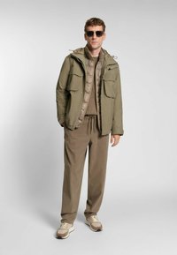 Giacca impermeabile verde oliva con diverse tasche, indossata sopra un gilet imbottito beige e una maglia lavorata marrone, abbinata a pantaloni larghi marroni e sneakers.